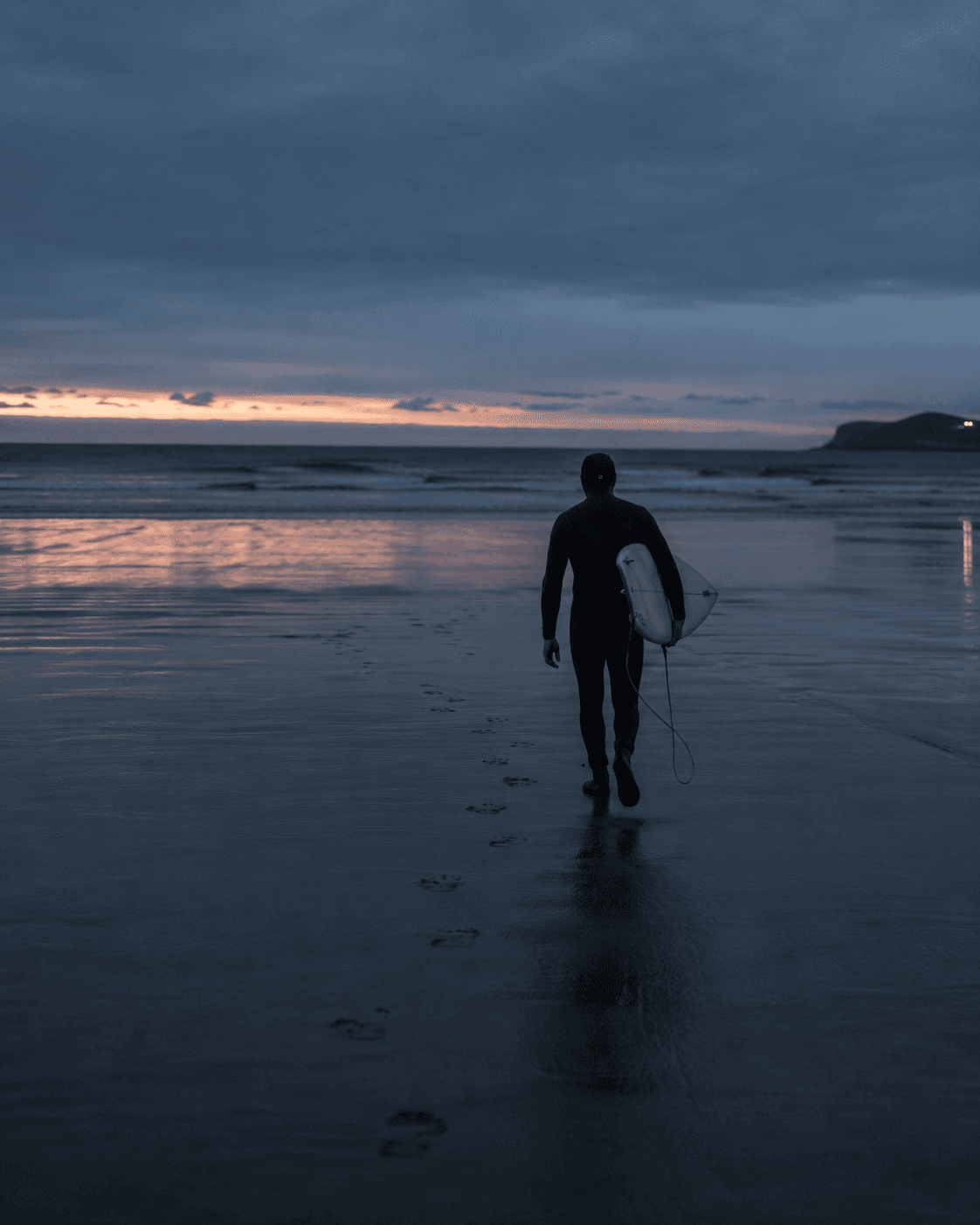 A surfer walking toward the ocean at dusk, board under arm, footprints trailing across wet sand.