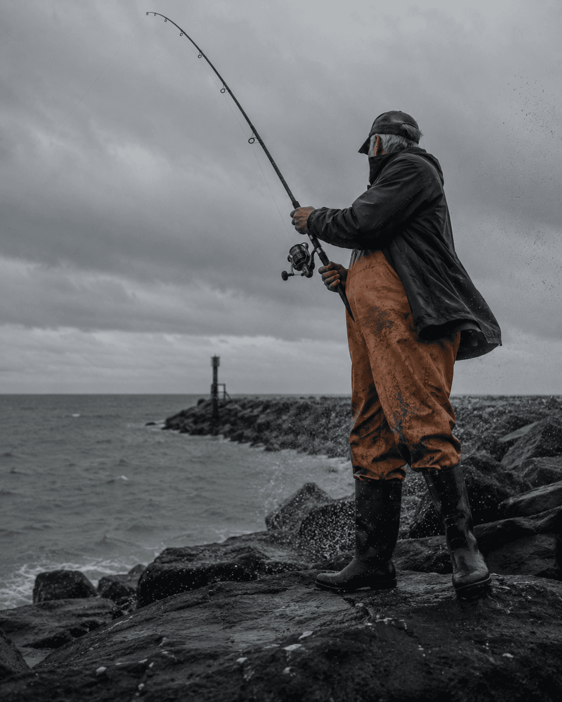 A fisherman casting from the jetty rocks in heavy rain, waders and rain gear against a grey sky.