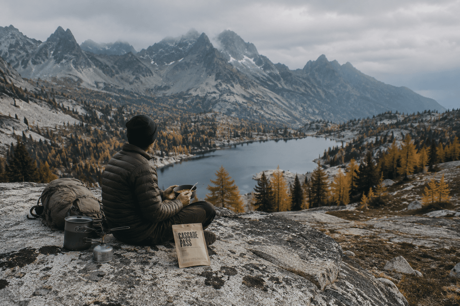 A hiker eating a meal on a rocky ridge overlooking an alpine lake.
