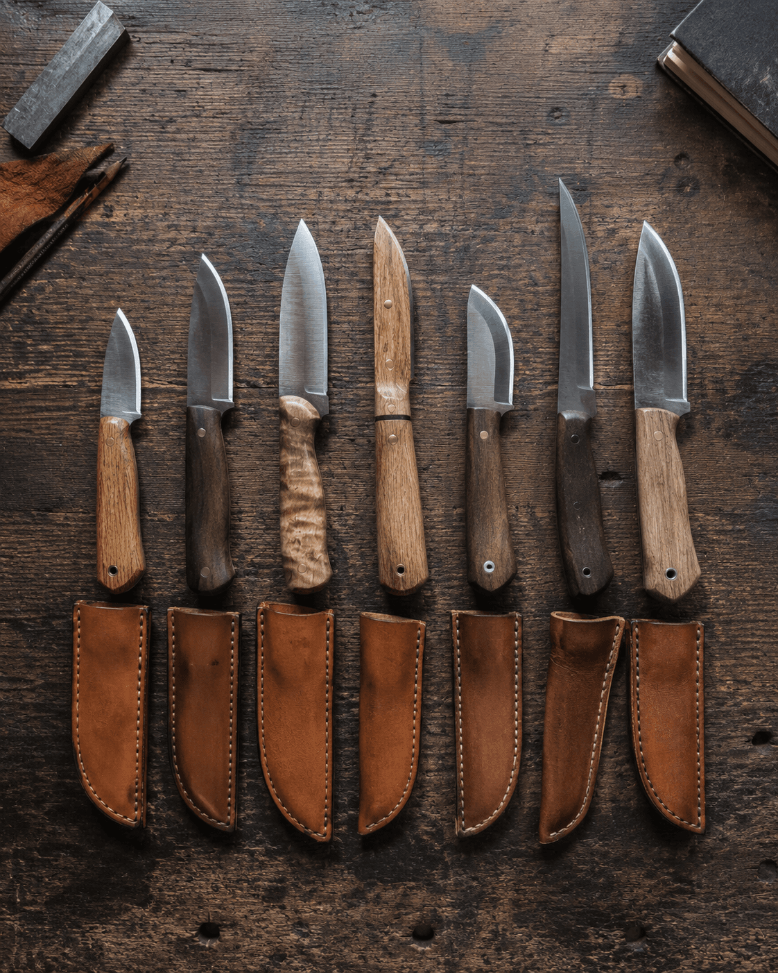 Seven finished Sandy River Forge knives laid out with their leather sheaths on a workbench