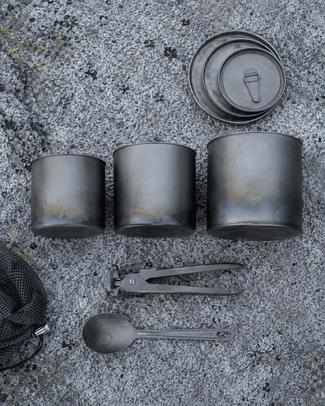 A flat lay of titanium cookware — three nesting cups, a lid, tongs, and a spoon on lichen-covered granite.
