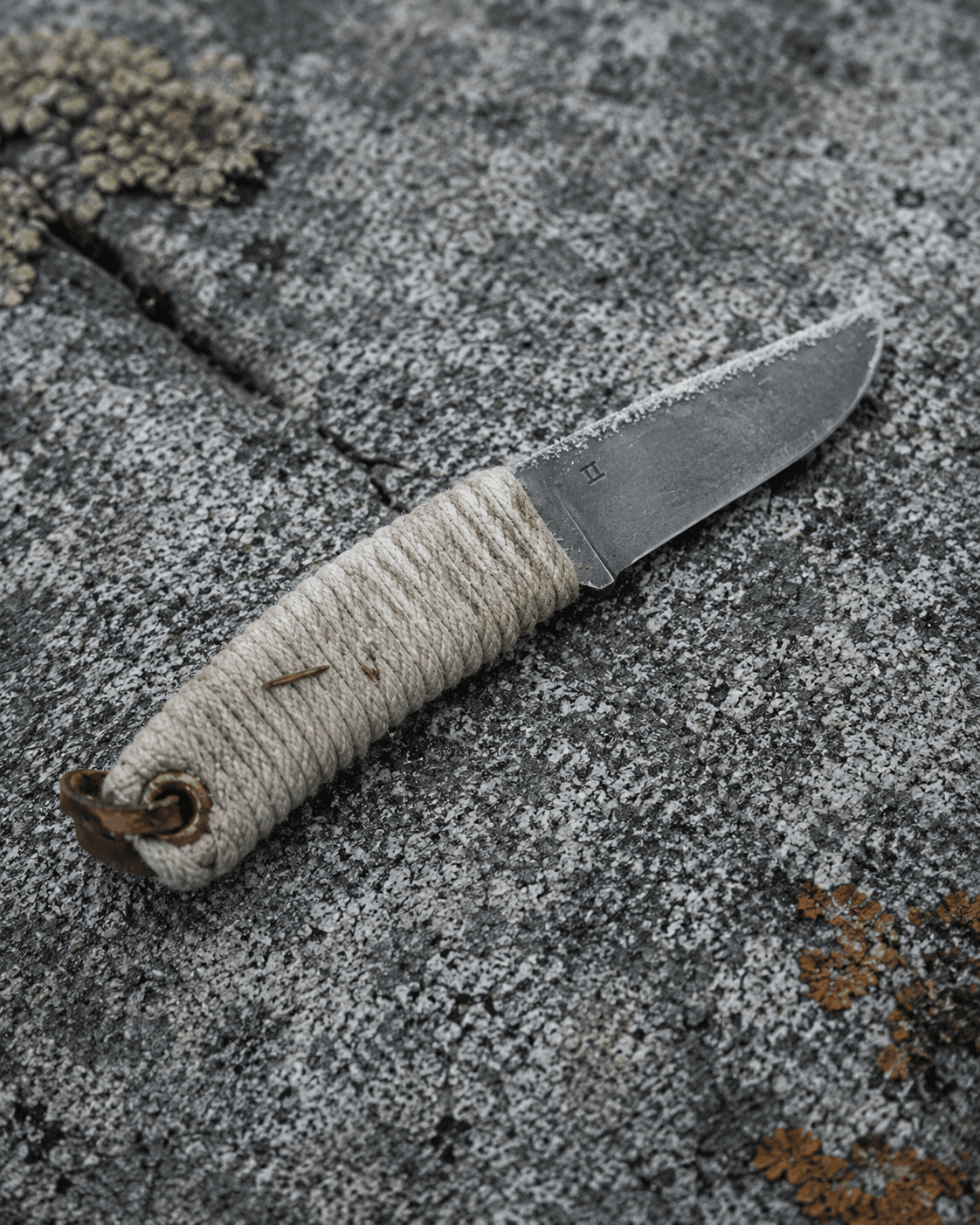 A hand-forged knife with a cord-wrapped handle resting on lichen-covered granite.