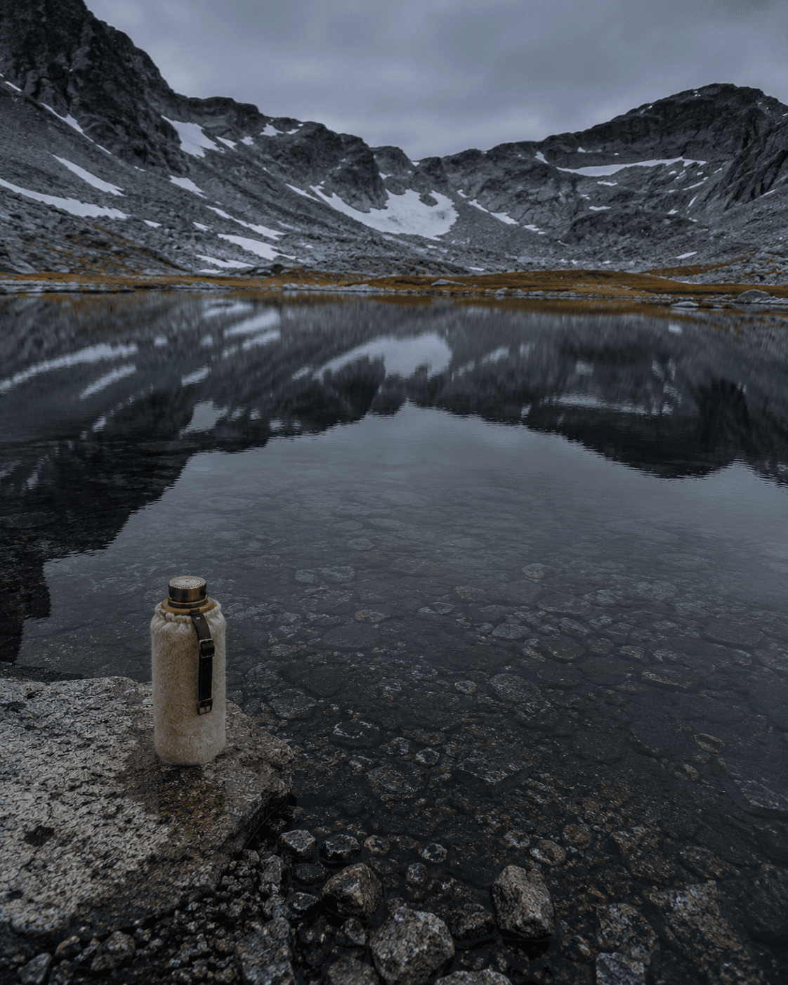A Tarn Goods water bottle on a rock at the edge of an alpine tarn, mountains reflected in the still water.